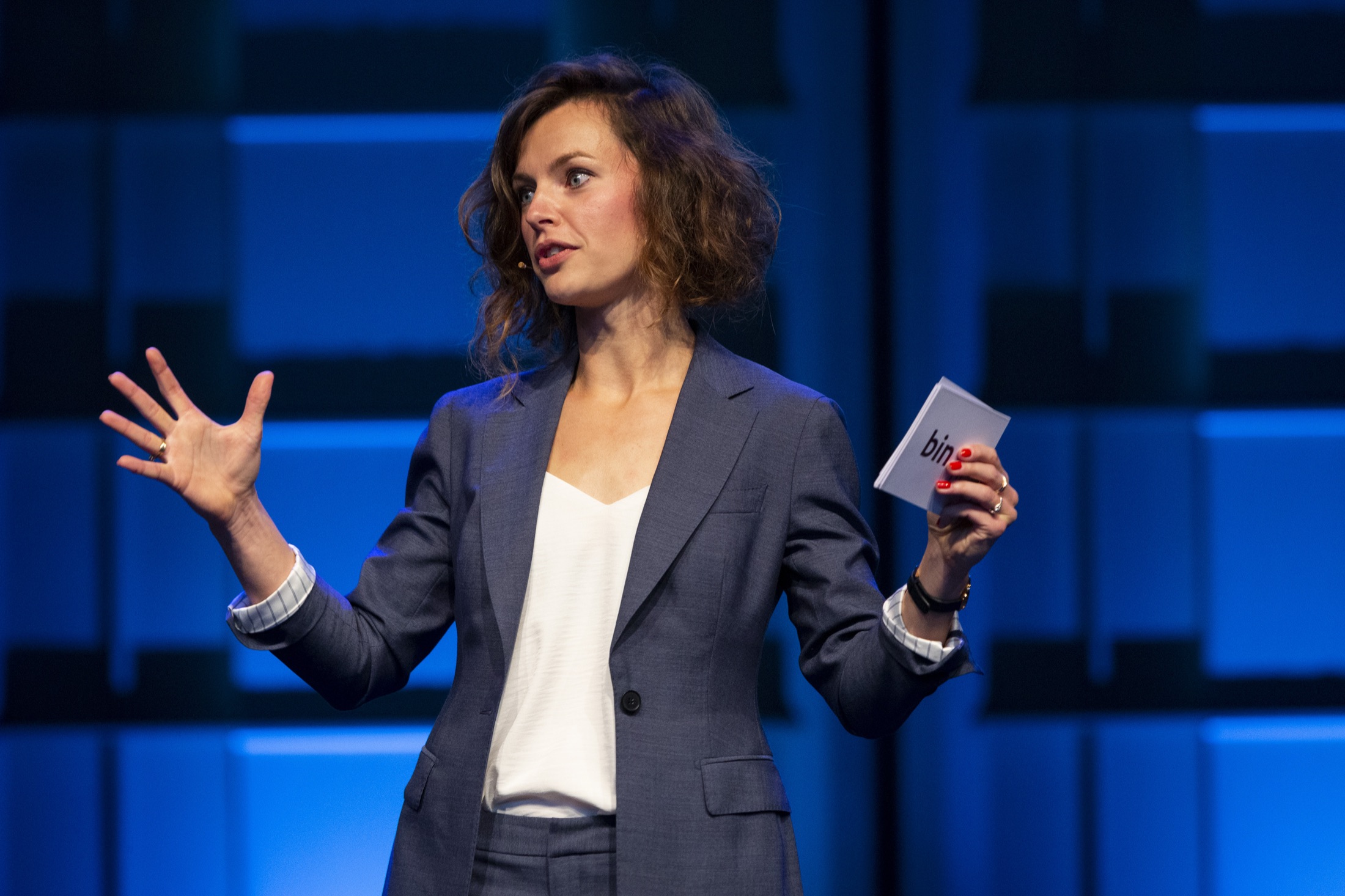 Liza Cornet spreekt op een podium, gesticulerend, tegen een blauw verlicht decor.