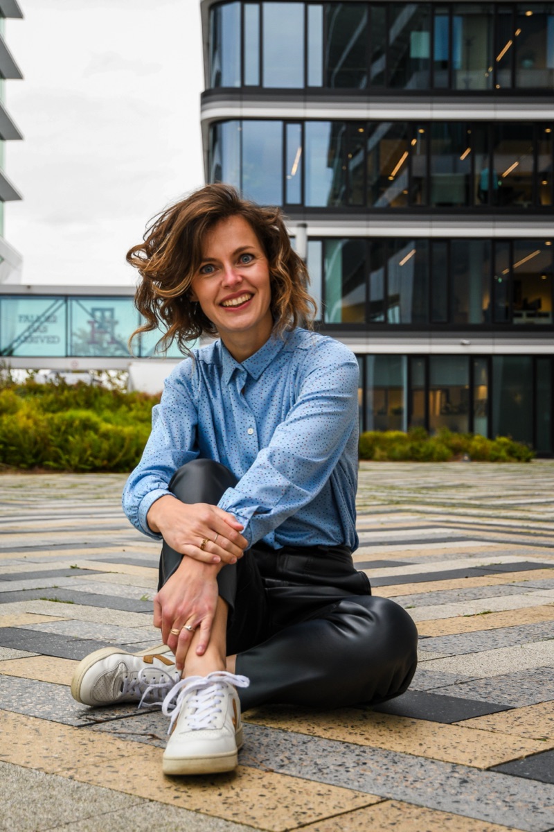 Portret van Liza Cornet, zittend op een plein voor een kantoorgebouw, lachend in de camera.