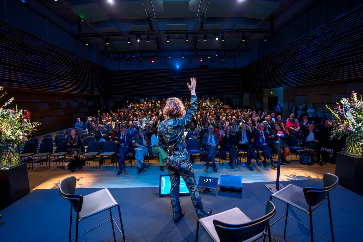 Liza Cornet op het podium met haar hand omhoog, tegenover een volle zaal waarin mensen hun hand opsteken.